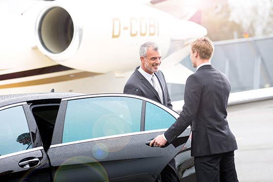 Ein Chauffeur öffnet einem Fahrgast die Tür einer Limousine auf dem Rollfeld eines Flughafens.